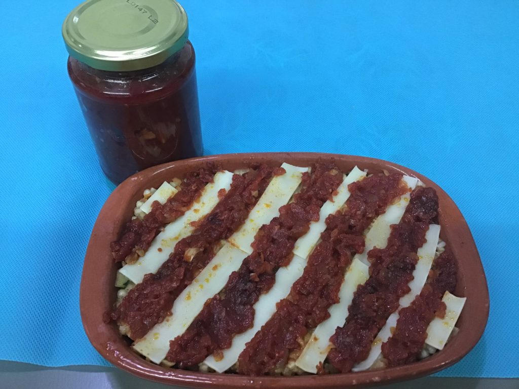 Un tarro de salsa de tomatate sin azúcar añadido preparado para conservar, delante una bandeja de barro que contiene una lasaña decorada con tiras de tomate y queso.