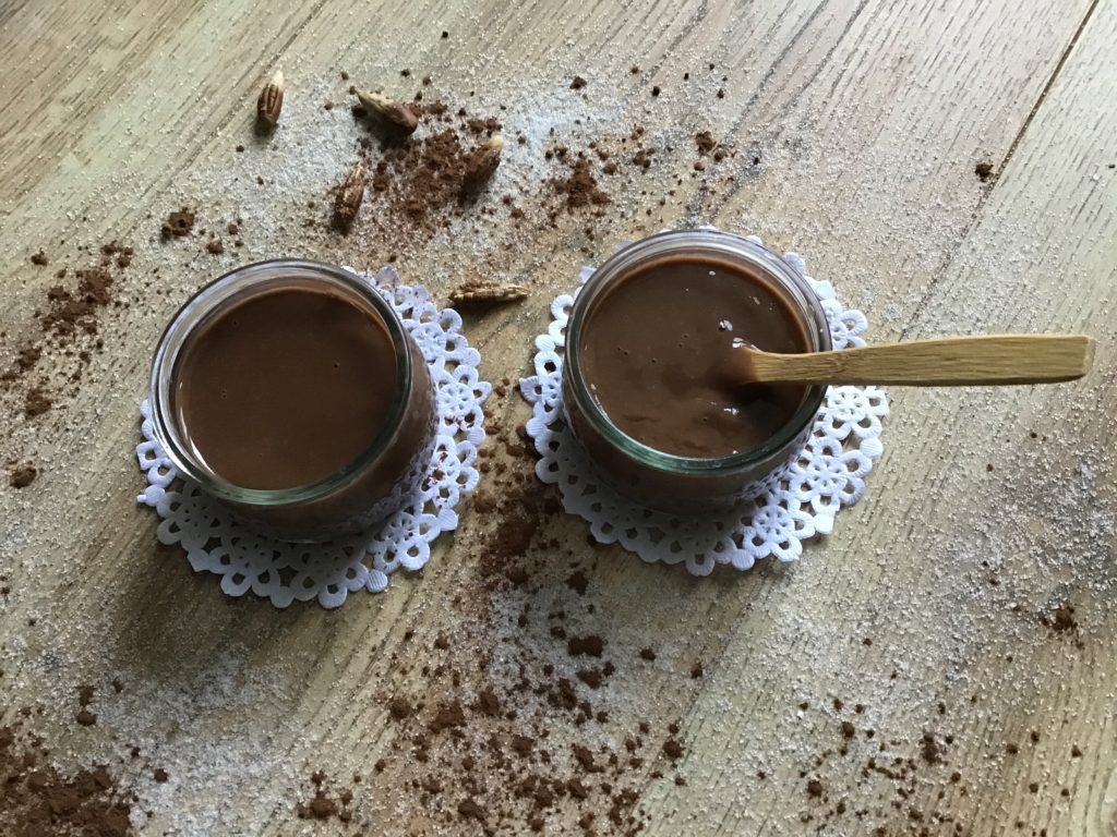 Dos basitos de natillas de chocolate sobre dos pequeñas blondas blancas. Al rededor hay derramado azúcar y café en polvo.