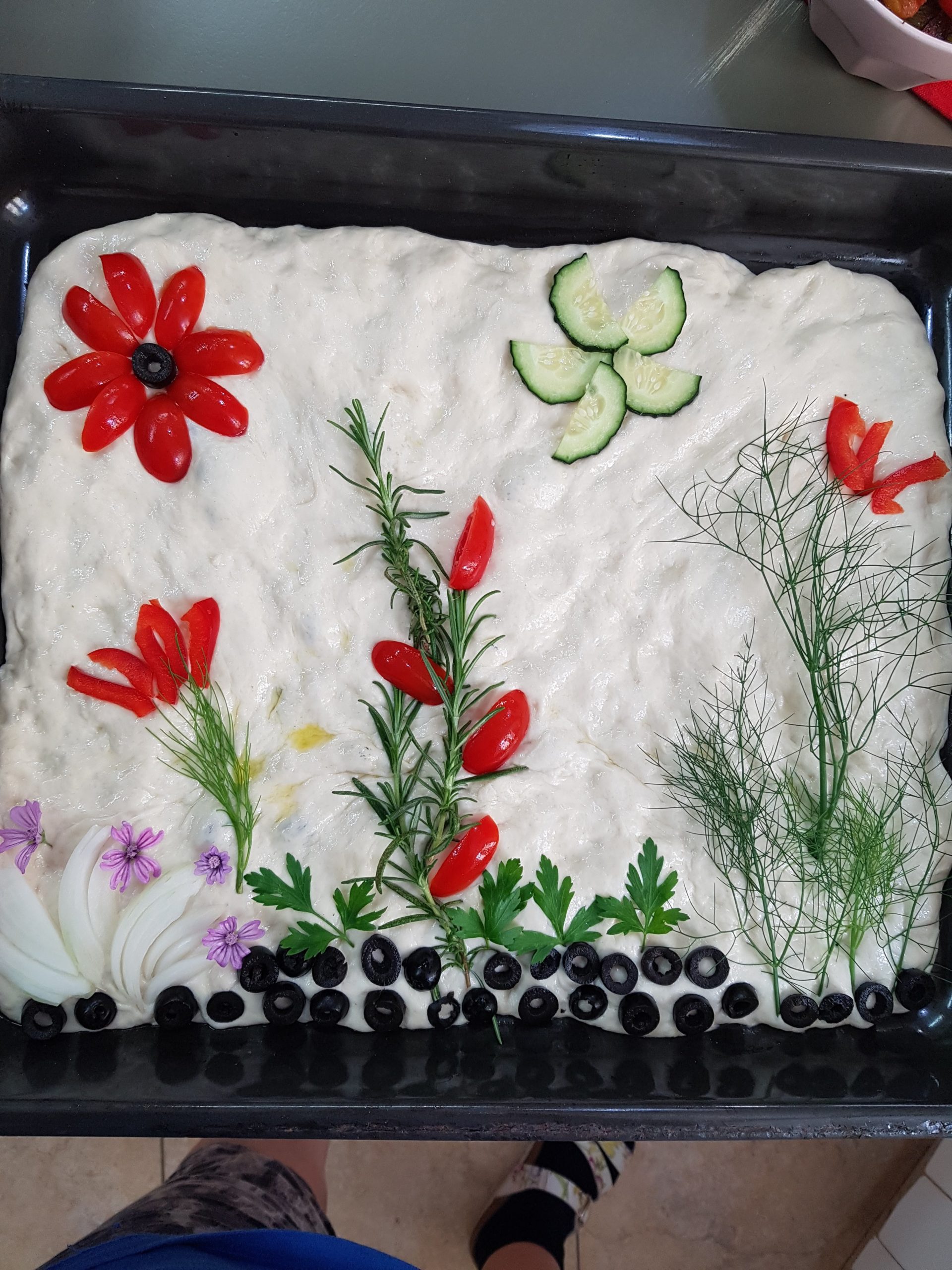 Esta imagen es de la decoración tan atractiva que se le puede dar a esta focaccia sin amasar para que sea espectacular. Hay tomatitos cherri cortados y tiras de pimiento rojo que simulan flores, también tallos de romero e hinojo y flores de malva perejil y algún trocito de pepino. La composición parece un pequeño jardín.