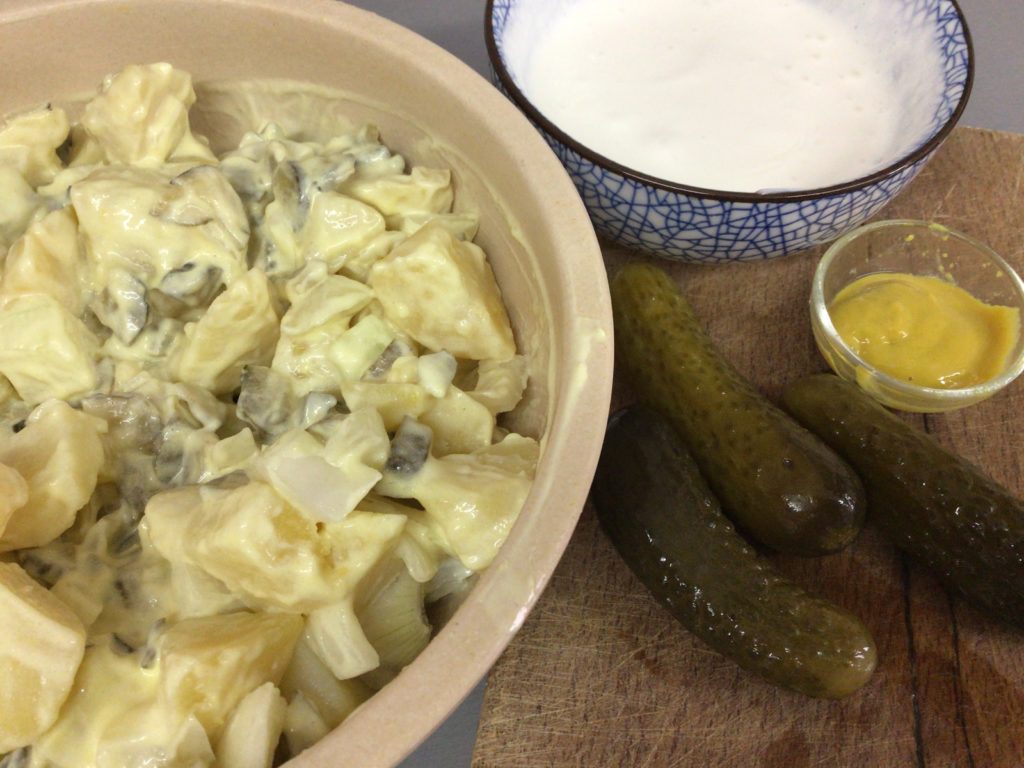 Una ensaladera llena de Kartoffelsalat, más un bol azul y blanco (con dibujo craquelado) con veganesa, un pequeño recipiente con mostaza de Dijon y tres pepinillos encurtidos que formarán parte de la ensalada alemana.