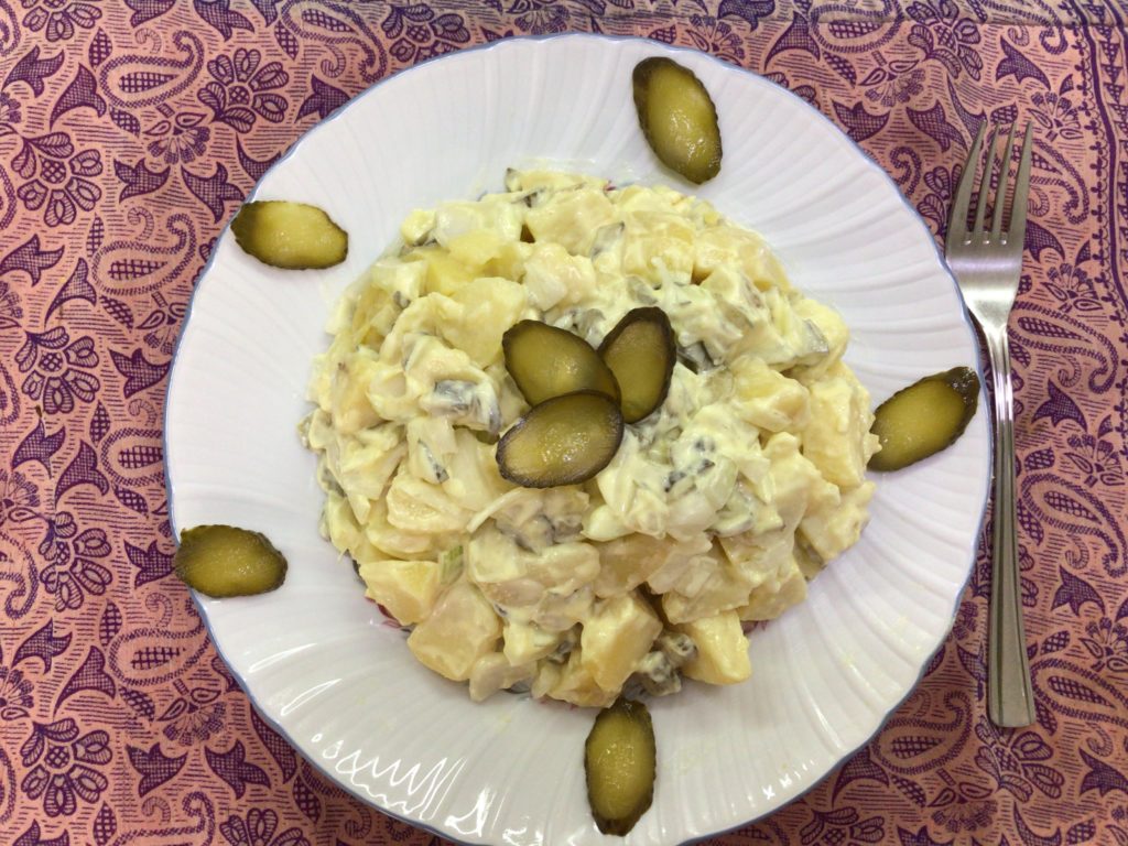 Ración de Kartoffelsalat servida en plato blanco y adornada con rebanadas de pepinillo.
