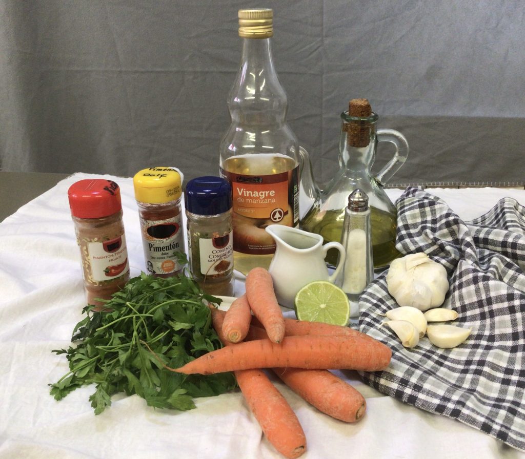 Esta imagen contiene todos los elementos para preparar esta ensalada marroquí de zanahorias: especias, limón, vinagre, ajos, sal, zanahorias y perejil fresco.